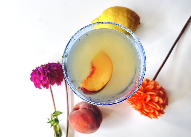 Cocktail overhead shot with peach slice in it. Peach and lemon on table with brightly colored mum flowers.